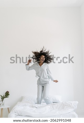 Vertical view of happy young afro american woman jumping up on bed, dance in white bedroom interior. Excited girl wearing in nightwear having fun at home, spend morning, listening music, smiling wide Royalty-Free Stock Photo #2062991048