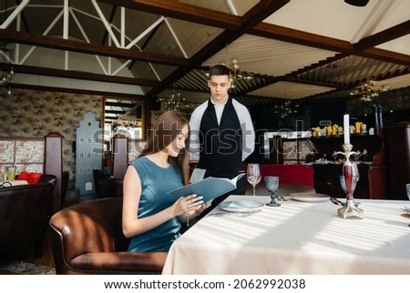 A young beautiful woman in a fine restaurant looks at the menu and makes an order to a young waiter in a stylish apron. Customer service.