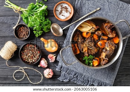 beef broth of beef meat on bones slow cooked with charred vegetables: carrot, onion, garlic, and spices served in a pot on a wooden background with ingredients, top view, flat lay