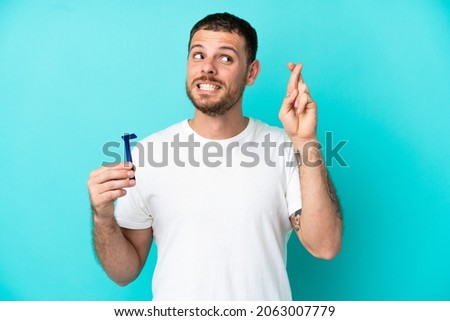 Brazilian man shaving his beard isolated on blue background with fingers crossing and wishing the best