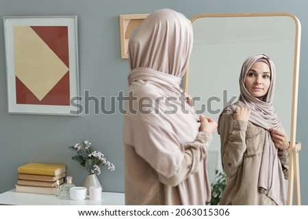 Young Muslim woman in hijab looking in large mirror while getting dressed for work in the morning