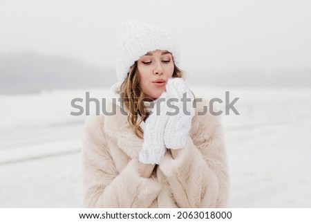 close up portrait of a young woman in a winter hat and white gloves blowing snow and warming her hand. Royalty-Free Stock Photo #2063018000