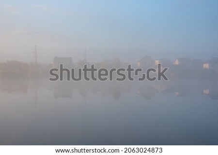Foggy misty landscape at the pond on early morning with small  buildings coming from the fog