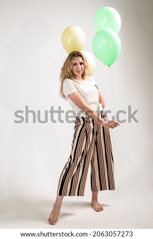 Funny girl, blonde, in a light T-shirt and striped trousers-skirt. a girl stands barefoot with balloons in her hands on a white background.