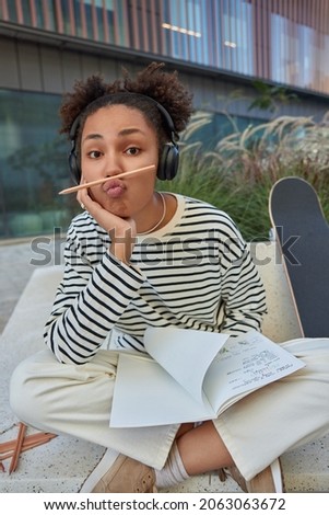 Funny curly haired teenage girl draws creative pictures in notebook keeps lips folded with pencil on sits crossed legs wears casual outfit rests after riding skateboard listens favorite music