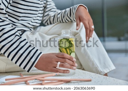 Faceless woman in casual clothes holds bottle of detox drink water with sliced lemon and cucumber rests aftr drawing picture surrounded by colored pencils. Healthy drinks sugar free beverage