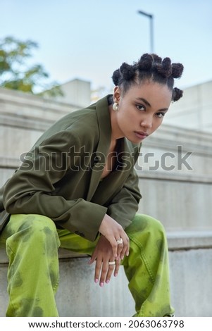 Cool teenage girl with stylish hairstyle makeup sits on steps outdoors wears fashionable green clothes earrings and rings looks self confident at camera belongs to youth subculture. Vertical shot