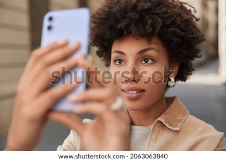 Close up shot of pretty young woman with curly hair makes photo on device shoots video via digital mobile phone strolls at street wears fashionable clothes focused at screen enjoys sunny day