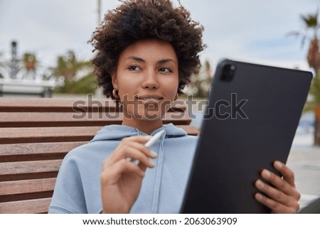 Dreamy satisfied woman with cutly hair holds modern tablet and stylus creats pictures or edits photos works outdoors wears casual hoodie poses at wooden bench duing daytime focused somewhere