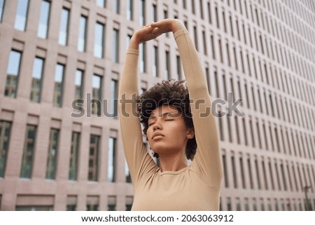Athletic calm woman with curly hair raises arms over head does stretching exercises wears beige tracksuit breathes deeply poses against modern building leads healthy lifestyle. Wellness concept Royalty-Free Stock Photo #2063063912