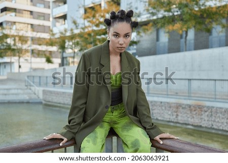 Beautiful female teenager looks seriously at camera during photo session dressed in stylish green clothes poses against scenic view outdoors has stroll at urban setting during nice sunny day