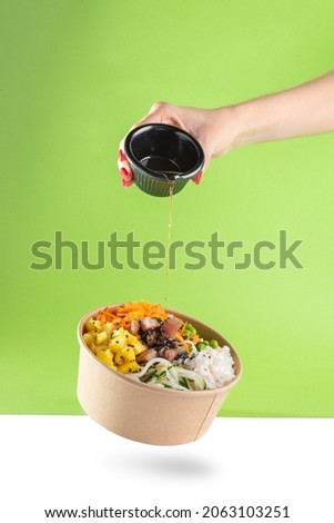 A woman's hand pours soy sauce into a floating bowl of salad poke and tuna, carrot, cucumber, and pineapple with a green background. white floor Royalty-Free Stock Photo #2063103251