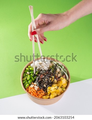 A woman's hand with chopsticks over a salad poke bowl and tuna, carrot, cucumber and pineapple with a green background. white floor Royalty-Free Stock Photo #2063103254