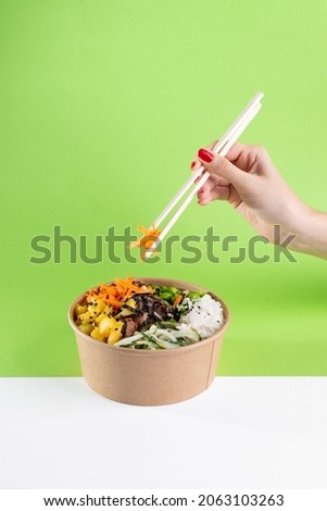 A woman's hand with chopsticks over a salad poke bowl and tuna, carrot, cucumber and pineapple with a green background. white floor Royalty-Free Stock Photo #2063103263