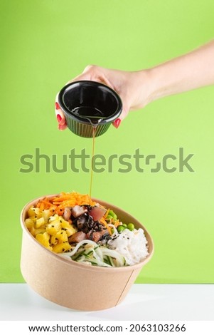A woman's hand pours soy sauce into a floating bowl of salad poke and tuna, carrot, cucumber, and pineapple with a green background. white floor Royalty-Free Stock Photo #2063103266
