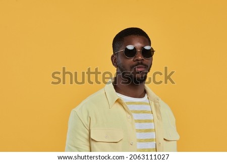 Minimal waist up portrait of trendy African-American man wearing sunglasses and looking at camera while standing against yellow background in studio, copy space