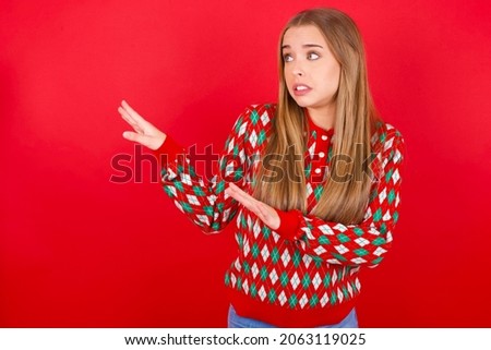 Displeased Young caucasian girl wearing Christmas sweater on red background keeps hands towards empty space and asks not come closer sees something unpleasant