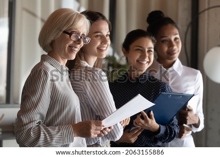 Motivated multiracial female team office workers of different generations look at one side smile ready to meet help client. Professional diverse businesswomen stand together hold documents clipboards Royalty-Free Stock Photo #2063155886