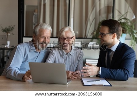 Smiling senior couple sit at lawyer broker designer office look at laptop listen to offer proposal of qualified specialist. Happy older spouses customers of male realtor choose flat house on pc screen Royalty-Free Stock Photo #2063156000