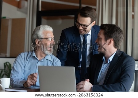 Intergenerational business team. Smiling male colleagues of diverse age have fun by computer at workplace. Older and younger men office employees gather at laptop talk laugh joke on funny work moment Royalty-Free Stock Photo #2063156024