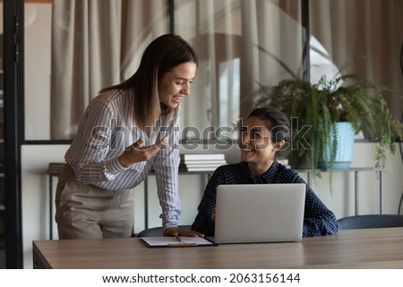 Two young females colleagues of diverse ethnicities discuss job chat hold pleasant conversation at office workplace. Young woman project manager assist indian coworker explain work in corporate app Royalty-Free Stock Photo #2063156144