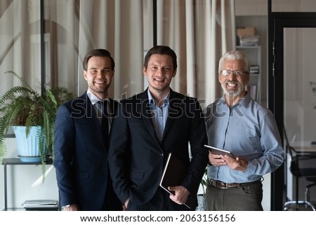 Smiling friendly group of three businessmen of different age private company staff posing for office portrait. Young male team leader ceo and two men aged and millennial subordinates looking at camera Royalty-Free Stock Photo #2063156156