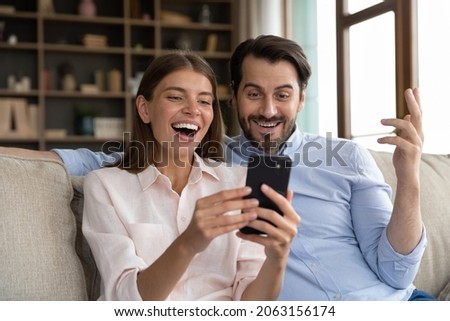 Overjoyed young family couple looking at telephone screen, feeling excited getting shopping discount sale deal offer, celebrating reading online lottery gambling win notification, sitting on sofa. Royalty-Free Stock Photo #2063156174