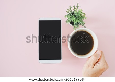 white mobile phone with clipping path on touchscreen and senior's hand holding a cup of hot coffee with blurred green fake small tree on sweet pink background