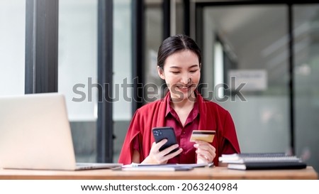 Beautiful young Asian businesswoman in red shirt smiling happy shopping online holding smartphone and credit card at office.