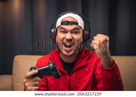 Playing video game. Young asian handsome man sitting on sofa holding joystick in living room. Happiness Streamer Indian man wearing headset playing game online in the darkroom.
