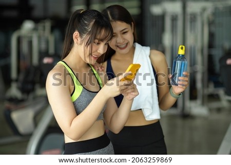 Two of sporty girl standing in fitness studio looking on smartphone screen together after finished workout.