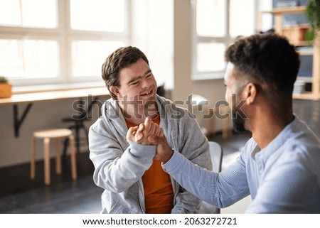Young happy man with Down syndrome with his mentoring friend celebrating success indoors at school. Royalty-Free Stock Photo #2063272721
