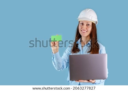 A female engineer in a hardcat with a laptop on a blue background, holding a green business card in her hands. Copyspace