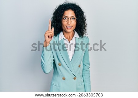 Young latin girl wearing business clothes and glasses pointing finger up with successful idea. exited and happy. number one. 