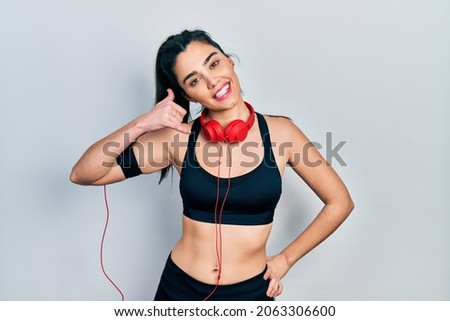 Young hispanic girl wearing gym clothes and using headphones smiling doing phone gesture with hand and fingers like talking on the telephone. communicating concepts. 