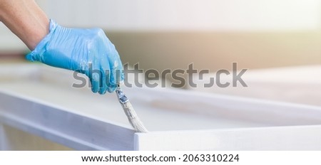 Male hands in blue gloves hold a paintbrush and paint a wooden board with white paint outdoors. The man is engaged in construction work and self-repair. Horizontal format banner. Close-up. Copy space