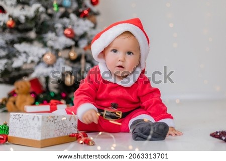 baby in a Santa costume is sitting next to the Christmas tree and playing with gift boxes. child opens a Christmas gift. winter new year's concept. space for text. High quality photo