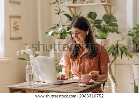 Positive young woman using a laptop and smartphone at home.Cozy home interior with indoor plants.Remote work, business,freelance,online shopping,e-learning,urban jungle concept.Selective focus. Royalty-Free Stock Photo #2063316752