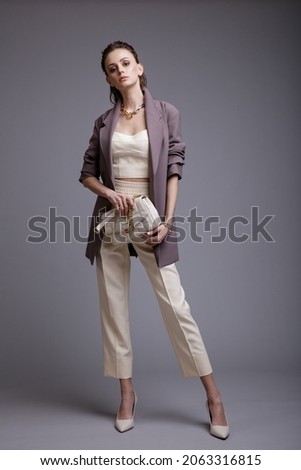 High fashion photo of a beautiful elegant young woman in a pretty purple jacket, white top, pants, accessories, handbag posing on gray background. Studio Shot. Slim figure. Make up, hairstyle