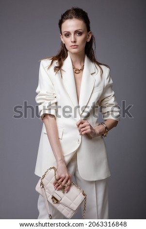 High fashion photo of a beautiful elegant young woman in a pretty white suit,  jacket, pants, trousers, accessories, handbag posing over gray background. Studio Shot. Slim figure. Make up, hairstyle.