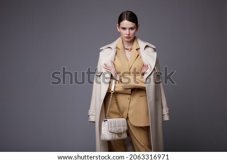 High fashion photo of a beautiful elegant young woman in a pretty white coat, beige sand suit, jacket, pants, trousers, accessories, handbag posing on gray background. Studio Shot. Slim figure. 