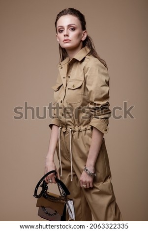 High fashion photo of a beautiful elegant young woman in a pretty beige sand jumpsuit, accessories, handbag posing on brown background. Studio Shot. Slim figure. Make up, hairstyle