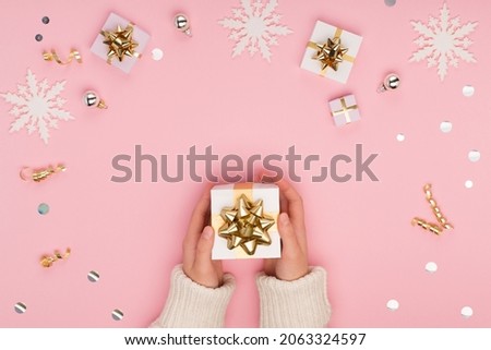 Woman hands holding white gift box on Christmas festive pastel pink background with gold and silver decorations, snowflakes. Holiday Xmas, winter, new year concept. Flat lay, top view, copy space.