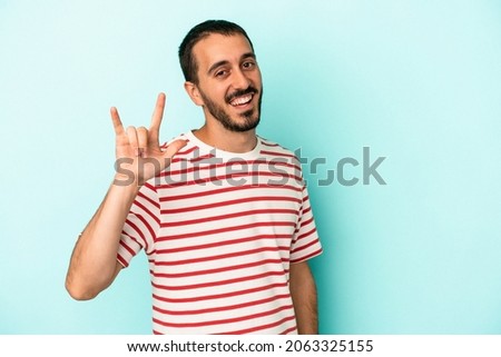 Young caucasian man isolated on blue background showing a horns gesture as a revolution concept.