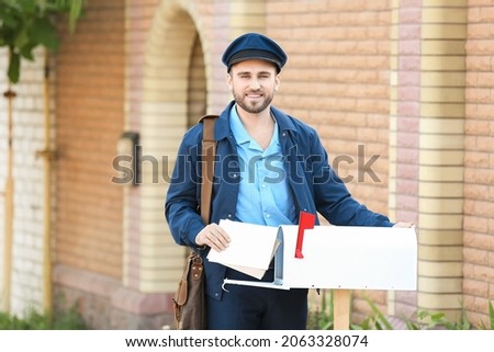 Handsome young postman putting letters in mail box outdoors Royalty-Free Stock Photo #2063328074