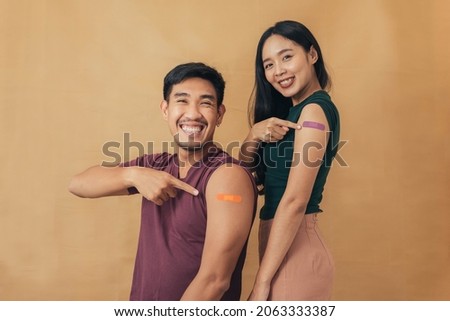 Asian man and woman showing shoulders after getting a vaccine. Happy couple showing arm with band-aids on after vaccine injection. Royalty-Free Stock Photo #2063333387