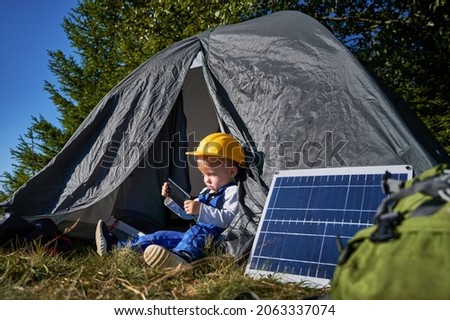 Young serviceman 3 years old resting near tent in camp and using cellphone technology for playing games during daytime, boy in uniform and helmet browsing website for watching online cartoons