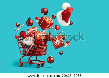Christmas composition with shopping cart with flying gifts, ornaments, candy canes and Santa cap on vibrant blue background. Creative Xmas or New year concept. Festive shopping or advertisement idea.