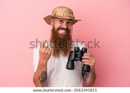 Young caucasian ginger man with long beard holding binoculars isolated on pink background pointing with finger at you as if inviting come closer.