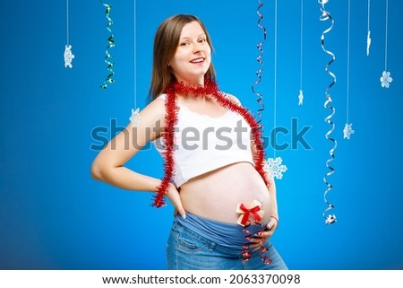 Pregnant woman with snowflakes on a blue background and red garland. Expecting a baby on Christmas eve.